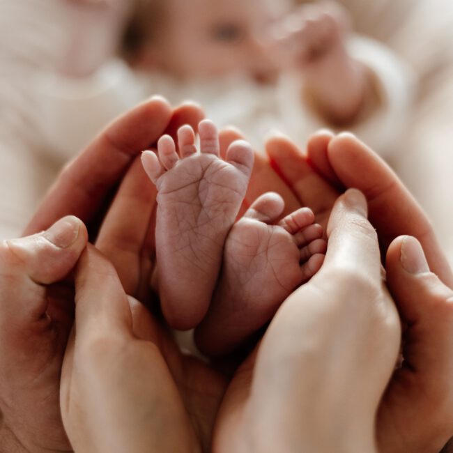 Eltern halten die kleinen Füße ihres Neugeborenen in den Händen, während das Baby im Hintergrund liegt.